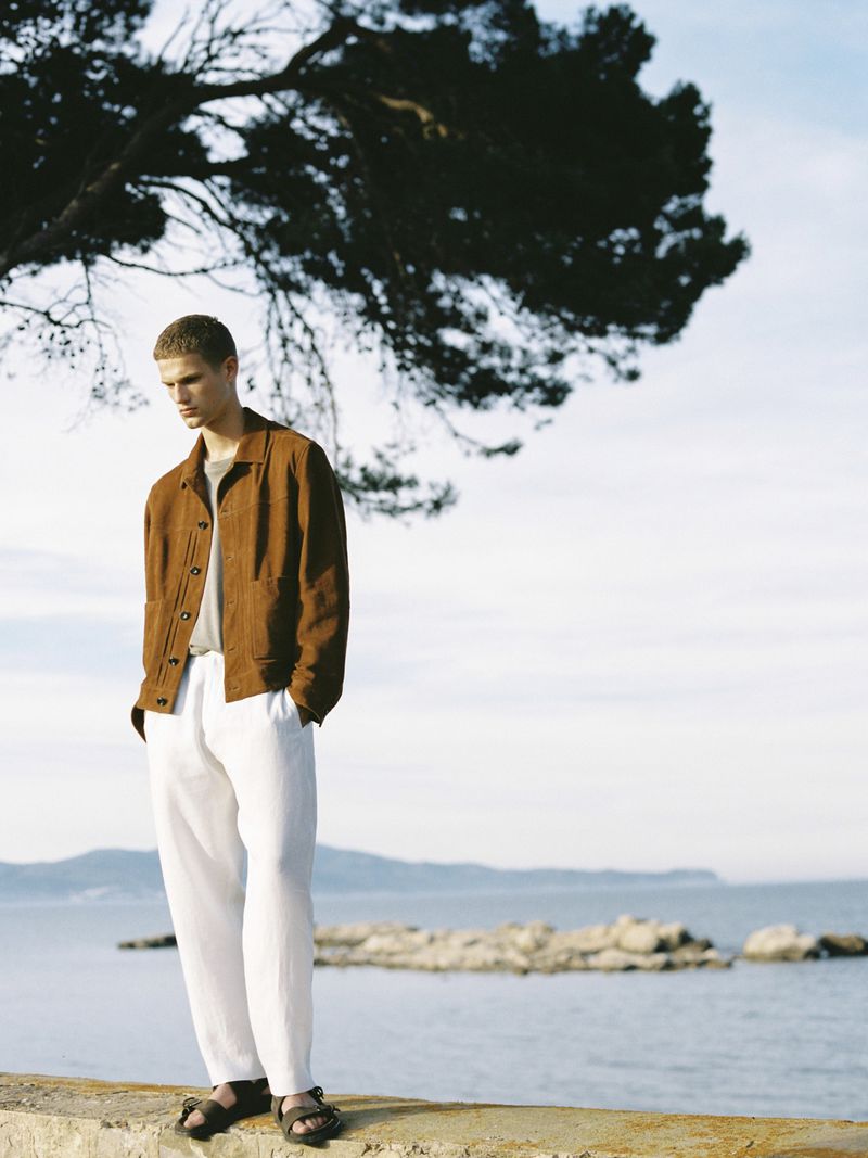 A young man stands on a concrete barrier near the ocean wearing a brown jacket, grey shirt and white pants, under a partly cloudy sky.
