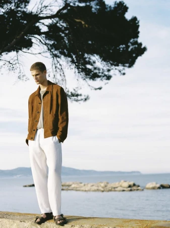 Teun van Stein wearing a brown jacket and relaxed white trousers standing on a stone ledge overlooking the sea.