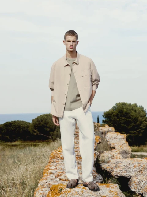 A fair-skinned male model poses outdoors wearing a tan shirt jacket, light green t-shirt, white pants, and sandals near the ocean.