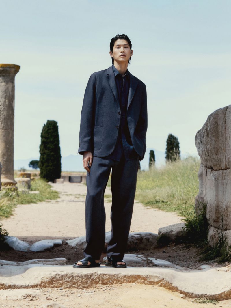 An Asian man in a dark blue suit stands on stone ruins with cypress trees in the background under a light blue daytime sky.