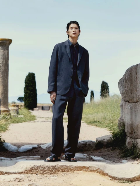 An Asian man in a dark blue suit stands on stone ruins with cypress trees in the background under a light blue daytime sky.