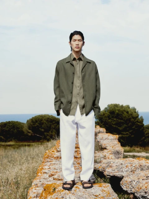 A full shot of an Asian male model wearing an olive green button-down jacket with white pants and sandals, standing on a stone structure.