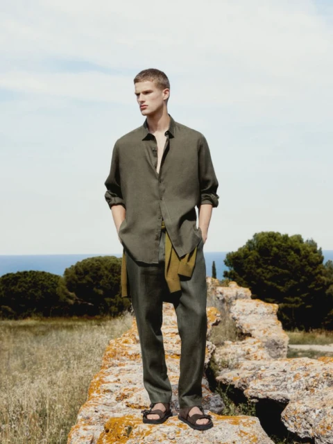 A full shot of a male model in an olive-green linen shirt and pants standing on a stone structure outdoors against a blue sky backdrop.