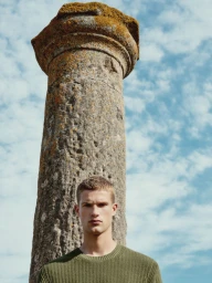 A male model wearing an olive green sweater stands before a tall stone column under a cloudy blue sky.