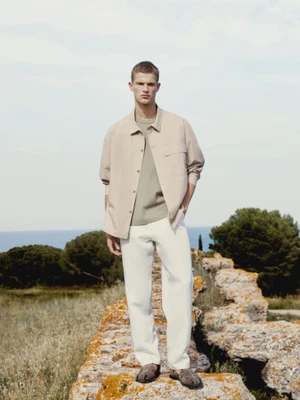 Male model in a beige overshirt and white trousers standing on a rocky ledge with the sea in the background.