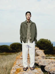 Model standing on a low stone wall wearing an olive jacket, button-down shirt, white trousers, and sandals against a coastal backdrop.