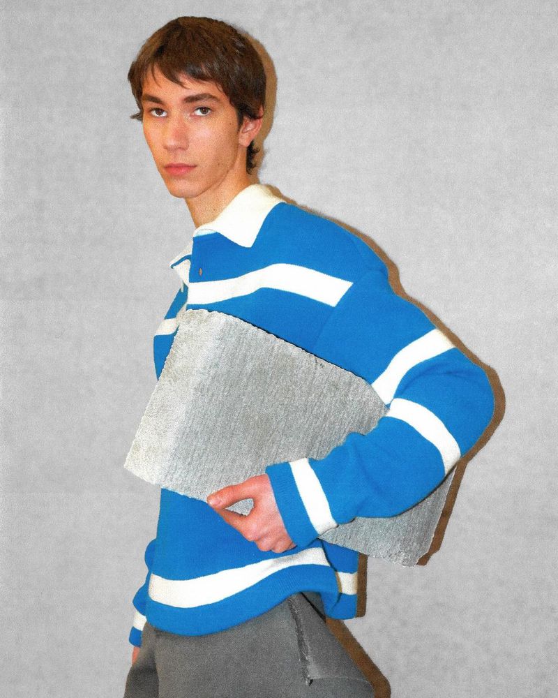 A young man models a blue and white striped rugby shirt, holding a textured gray object, posed in a neutral indoor setting.