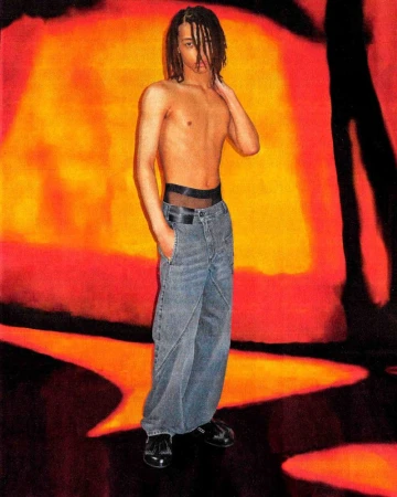 A young, shirtless person with dreadlocks is standing in a pair of baggy jeans in front of an orange and red background.