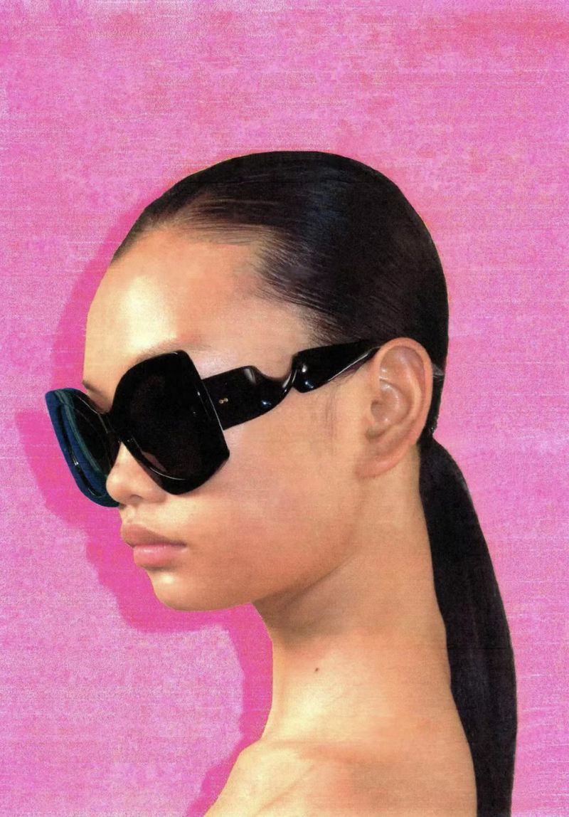 A woman with dark hair pulled back, wearing black sunglasses, posing in a studio with a pink textured background. Beauty shot, fashion.