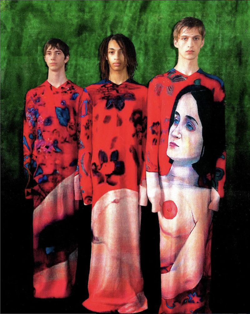 Four models in artful, surreal tunics stand against a green mottled backdrop, creating a fashion editorial with a fine art sensibility.
