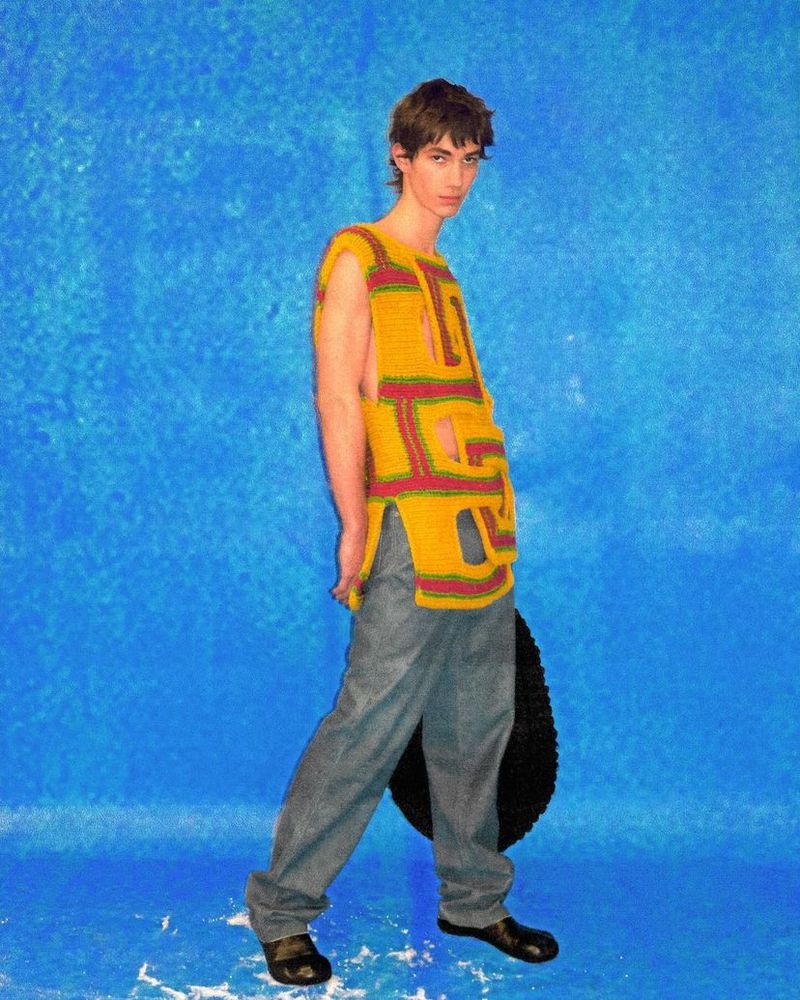 A male model poses in a yellow knit top and jeans against a blue mottled backdrop, with a large black wheel propped behind him.