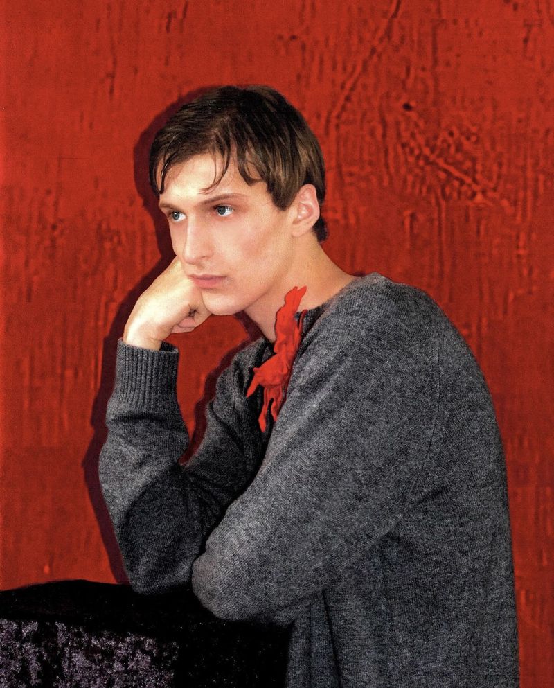 Portrait of a young adult male in a grey sweater, resting his chin on his hand, set against a textured red background with soft, diffused lighting.