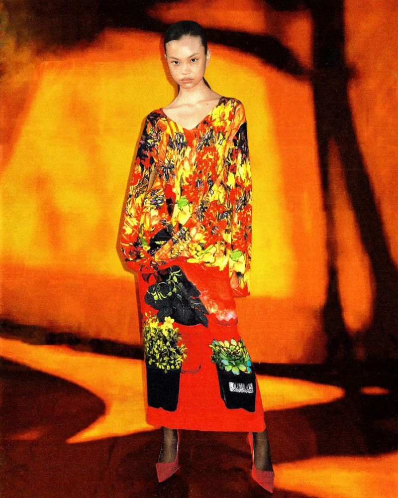 An Asian woman stands in a brightly lit room wearing a floral print top and a red skirt with plant pot designs, matched with red high heels.