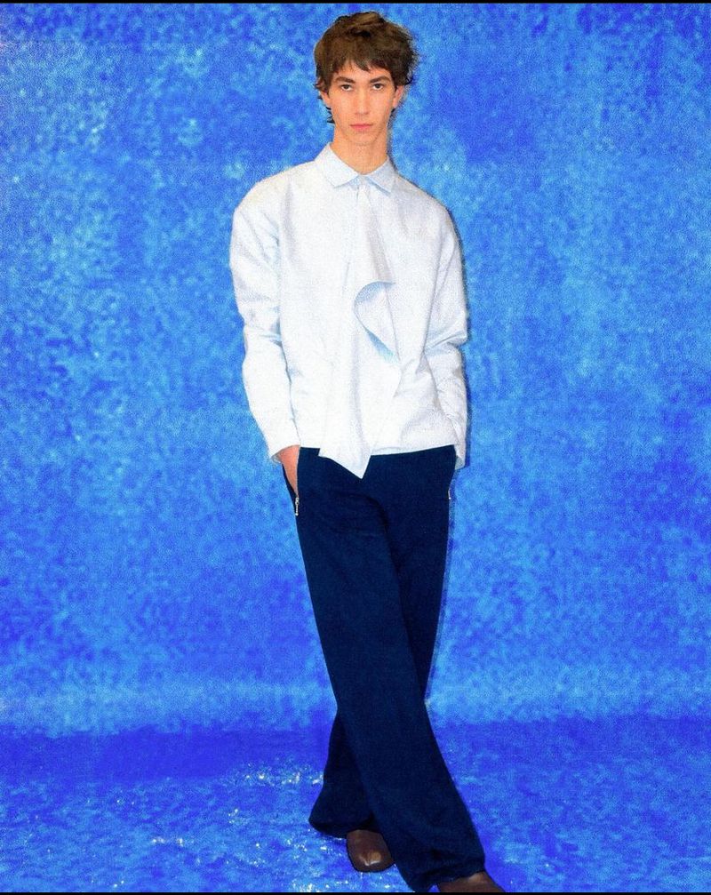 A young adult stands in front of a mottled blue background, wearing a light blue button-down shirt, dark blue pants, and brown shoes.