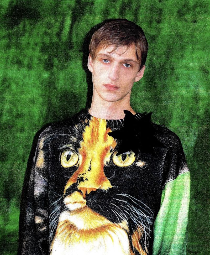Indoor portrait of a fair-skinned young adult male with light brown hair wearing a sweater with a large calico cat face graphic.