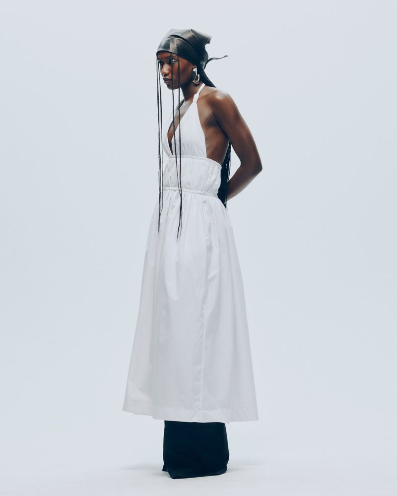 Fashion photo features a Black woman in a white dress and black head wrap against a pale background, showcasing minimalist fashion.