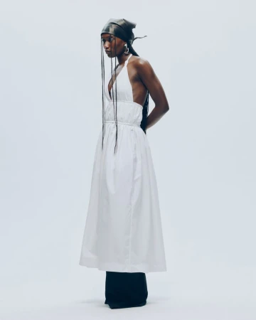 Fashion photo features a Black woman in a white dress and black head wrap against a pale background, showcasing minimalist fashion.