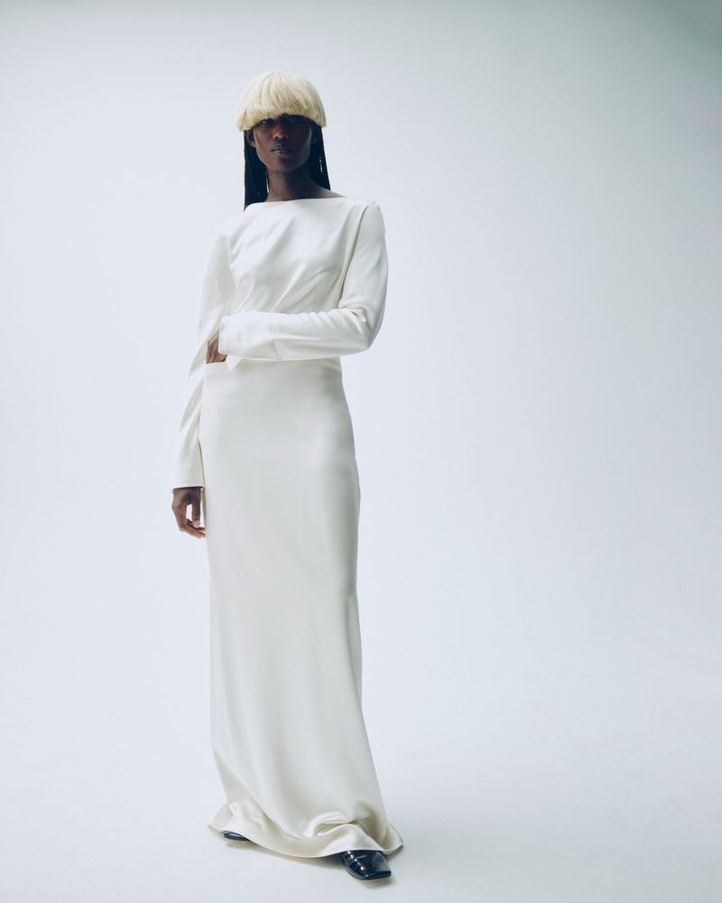 A Black woman stands in a white floor-length dress and blond wig against a white backdrop in a fashion-style shot.