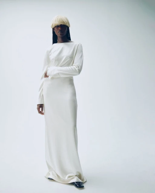 A Black woman stands in a white floor-length dress and blond wig against a white backdrop in a fashion-style shot.