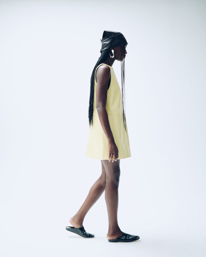 A woman in a yellow dress and black head covering walks against a bright white background, showcasing a fashion-forward, minimalist style.