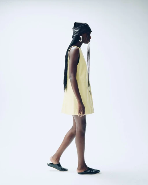 A woman in a yellow dress and black head covering walks against a bright white background, showcasing a fashion-forward, minimalist style.