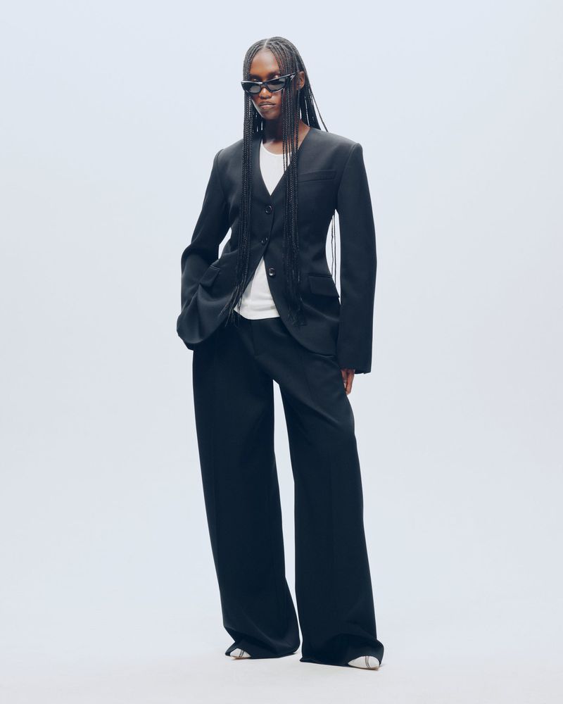 Model Essence Taylor wearing a tailored black collarless blazer, wide-leg trousers, and sunglasses against a white studio background.