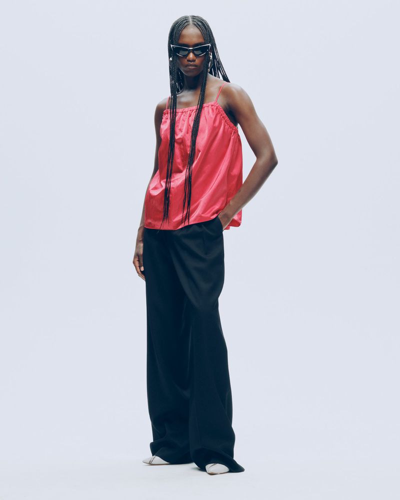 Full-length studio shot of a Black woman wearing a pink camisole, black wide-leg pants, and sunglasses against a light gray backdrop.