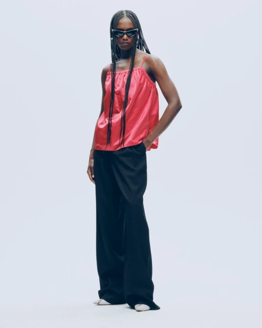 Full-length studio shot of a Black woman wearing a pink camisole, black wide-leg pants, and sunglasses against a light gray backdrop.