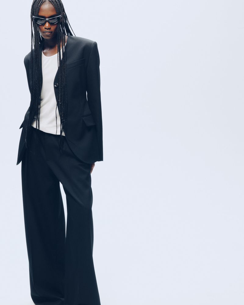 Essence Taylor wearing a black tailored blazer, wide-leg trousers, white t-shirt, and sunglasses against a white studio background.
