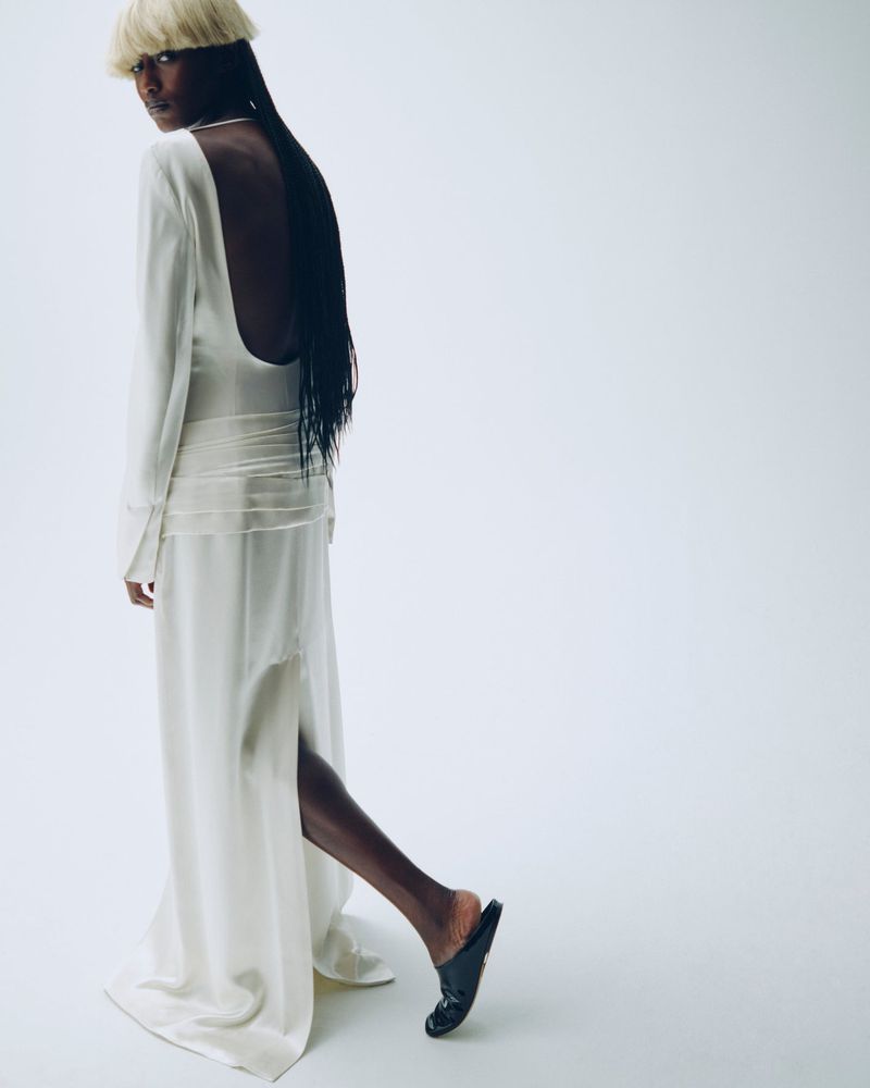 Dark-skinned female fashion model in white, long-sleeved, open-back dress with blonde bowl cut, posing in front of a bright white background.