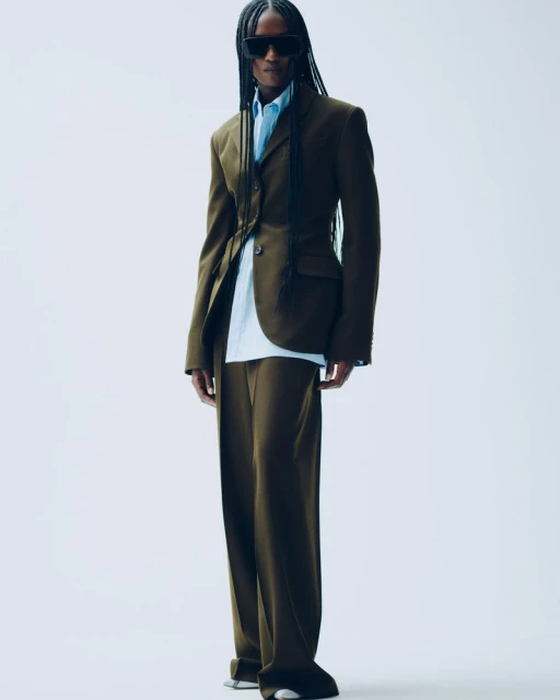 Stylish adult in tailored olive suit with braids and sunglasses standing in a neutral studio. Fashion photography, modern tailoring.