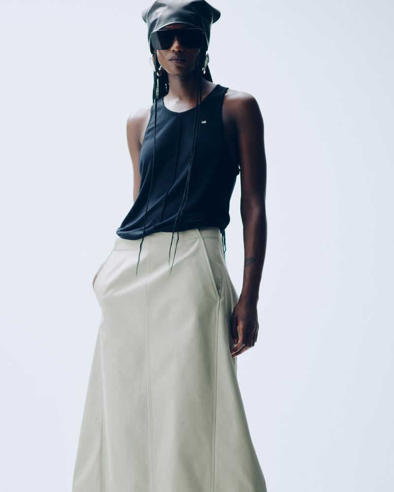 A fashionable Black woman with braids, wearing a black tank top and a beige maxi skirt, poses against a stark white background in a studio.