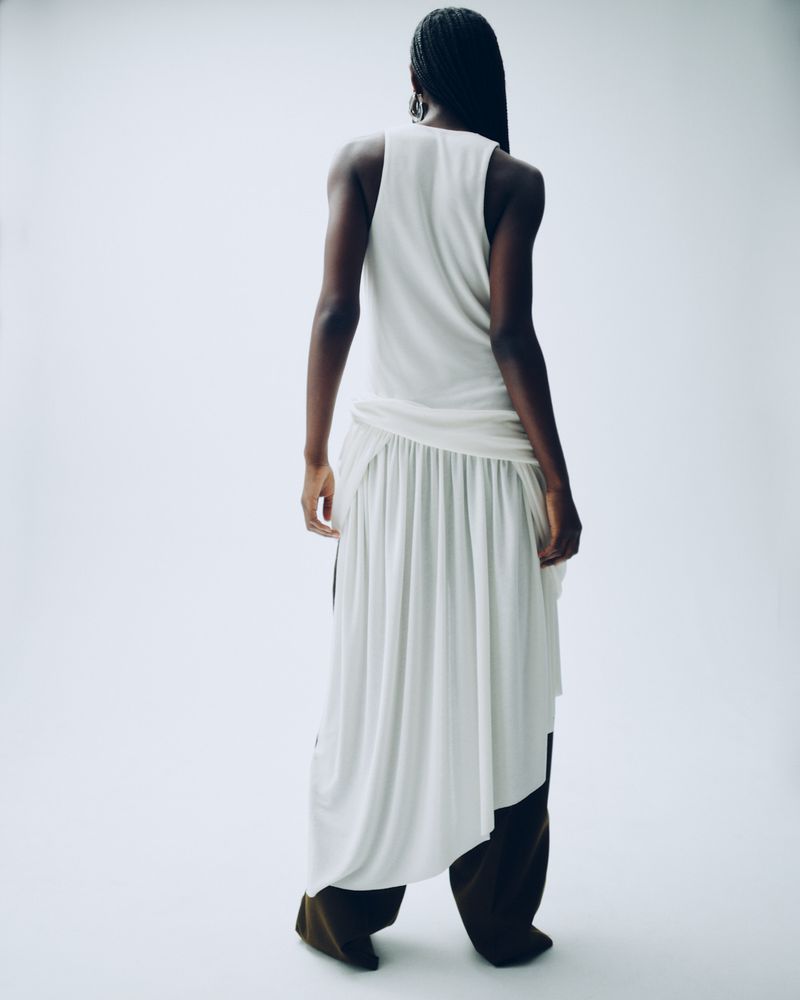 Back view of an adult woman in a white top and wide-legged brown pants standing in a white photo studio wearing neutral fashion clothing.