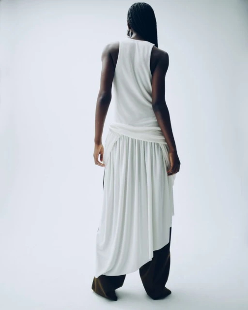 Back view of an adult woman in a white top and wide-legged brown pants standing in a white photo studio wearing neutral fashion clothing.