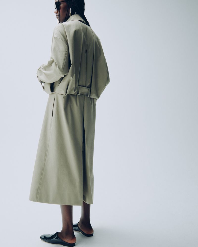 Fashionable adult female wearing a beige skirt and jacket, along with sunglasses and black flats, posing in a photo studio.