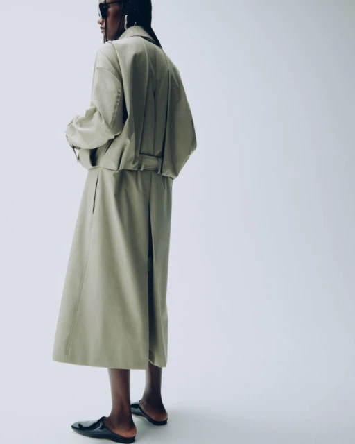 Fashionable adult female wearing a beige skirt and jacket, along with sunglasses and black flats, posing in a photo studio.
