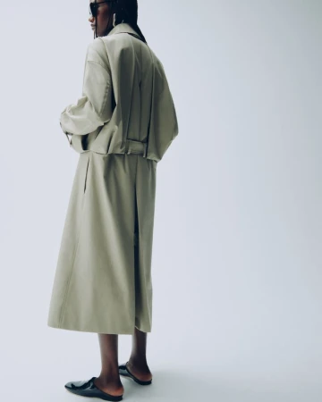 Fashionable adult female wearing a beige skirt and jacket, along with sunglasses and black flats, posing in a photo studio.