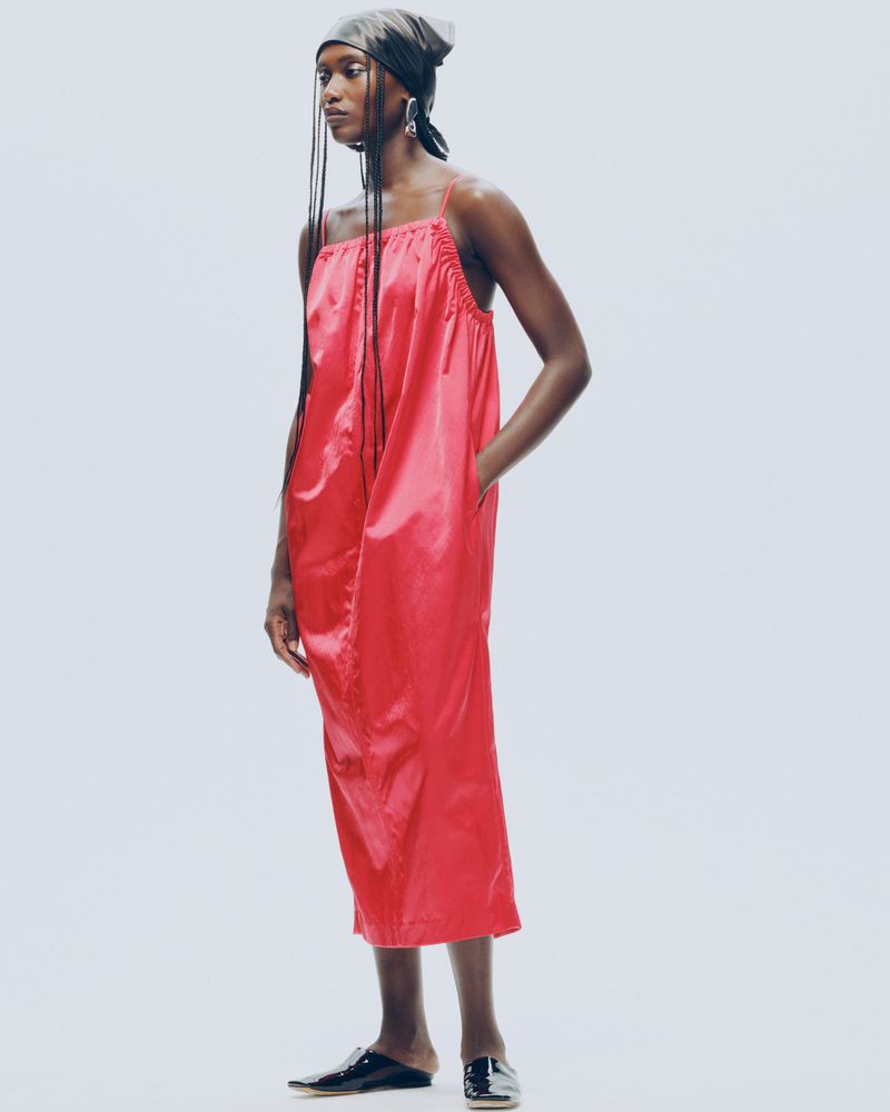 Full-length portrait of a Black woman in a shiny red dress and headscarf against a bright background, showcasing contemporary fashion.