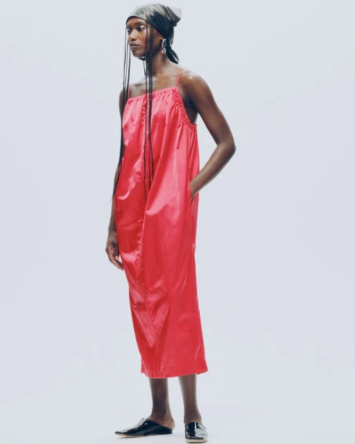 Full-length portrait of a Black woman in a shiny red dress and headscarf against a bright background, showcasing contemporary fashion.