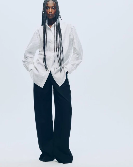 Fashionable woman in a white striped shirt and dark trousers against a white backdrop. Long dark braids frame her face.