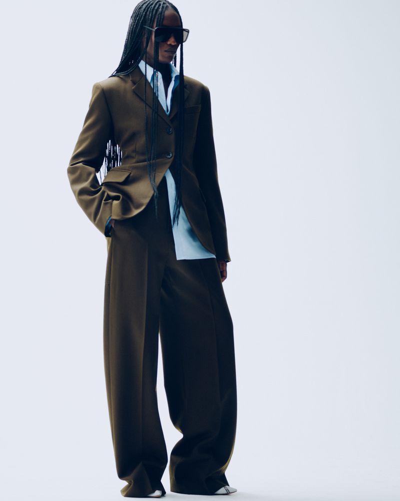 A woman in a brown suit with large, wide-legged pants poses in a bright photo studio, looking confidently forward and wearing dark sunglasses.