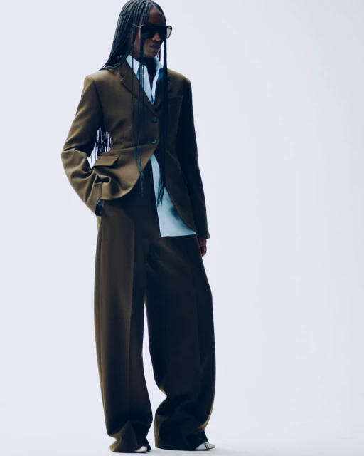 A woman in a brown suit with large, wide-legged pants poses in a bright photo studio, looking confidently forward and wearing dark sunglasses.