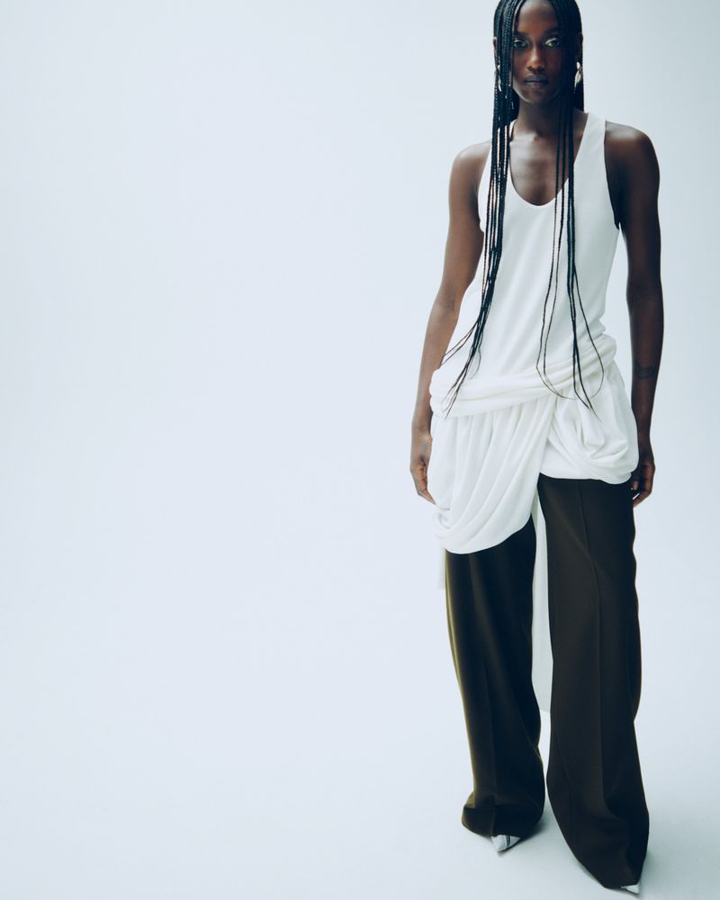 A fashionable Black woman poses in a draped white top and olive-green pants against a bright white backdrop in a studio fashion shot.