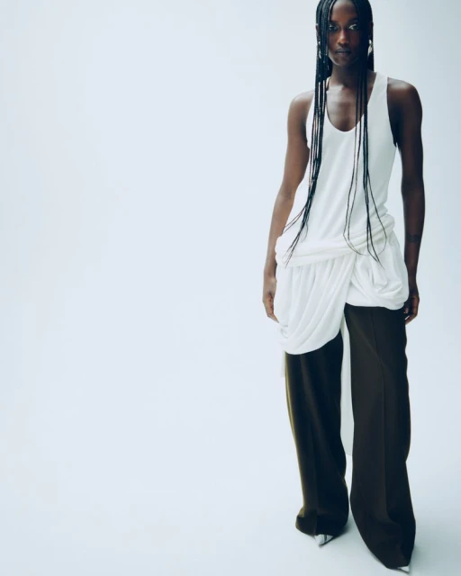 A fashionable Black woman poses in a draped white top and olive-green pants against a bright white backdrop in a studio fashion shot.