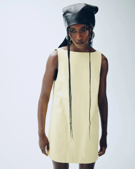 Fashion shot of a single African-descent adult female model wearing a pale yellow mini-dress with a black leather head covering.