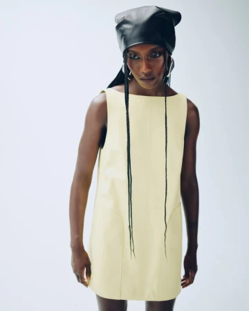 Fashion shot of a single African-descent adult female model wearing a pale yellow mini-dress with a black leather head covering.