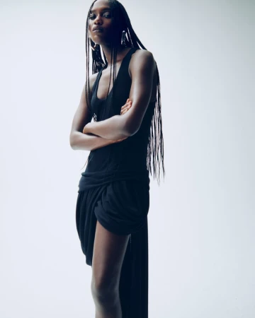 Model Essence Taylor wearing a draped sleeveless black dress with long braids against a white studio background.