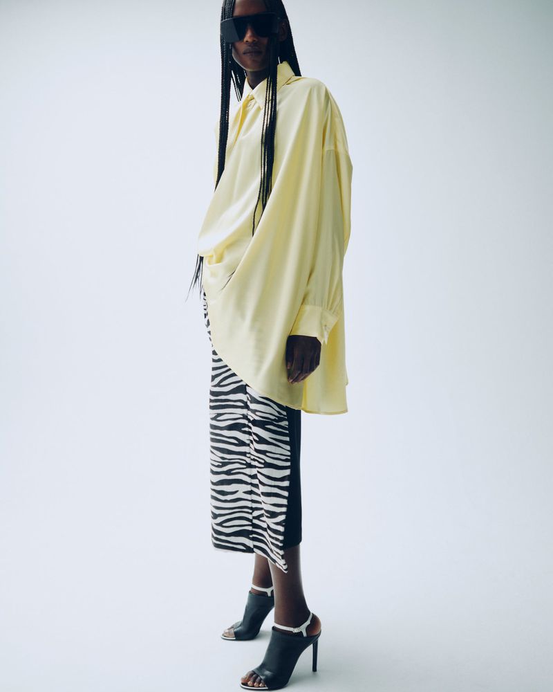 Fashionable woman posing in oversized yellow shirt and zebra-print skirt with stiletto heels in a light-filled studio setting.