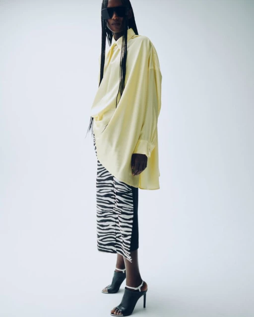 Fashionable woman posing in oversized yellow shirt and zebra-print skirt with stiletto heels in a light-filled studio setting.