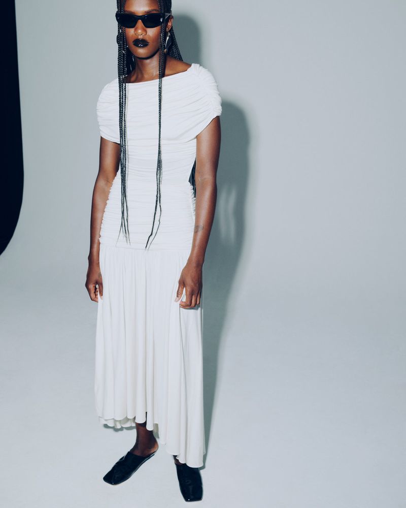 An African American woman models a white dress and black flats in a studio. She wears black sunglasses, and her braids hang long.